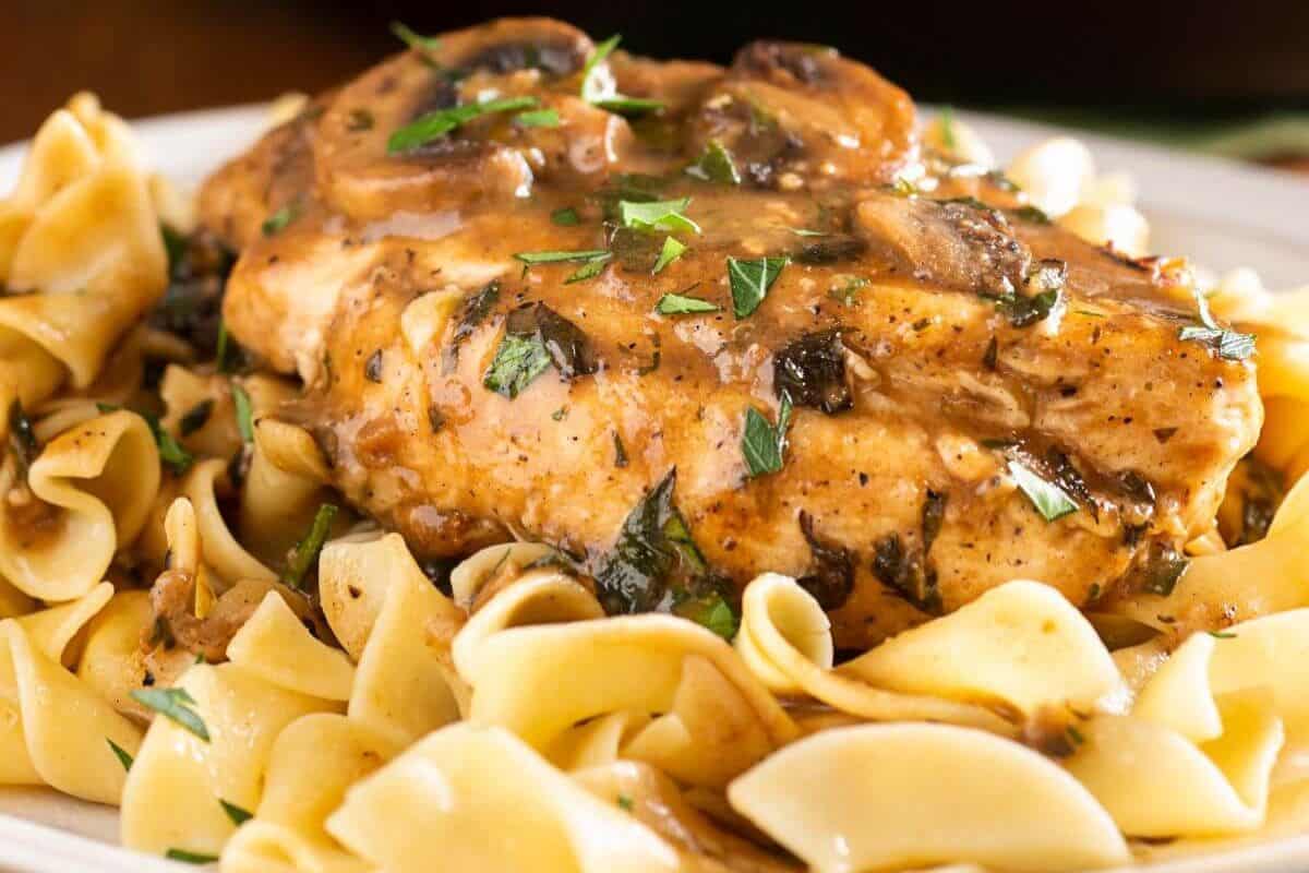 A close-up of a plate of Chicken Marsala. A golden-brown chicken breast is topped with a rich brown sauce featuring sliced mushrooms and herbs, and it rests on a bed of wide egg noodles.