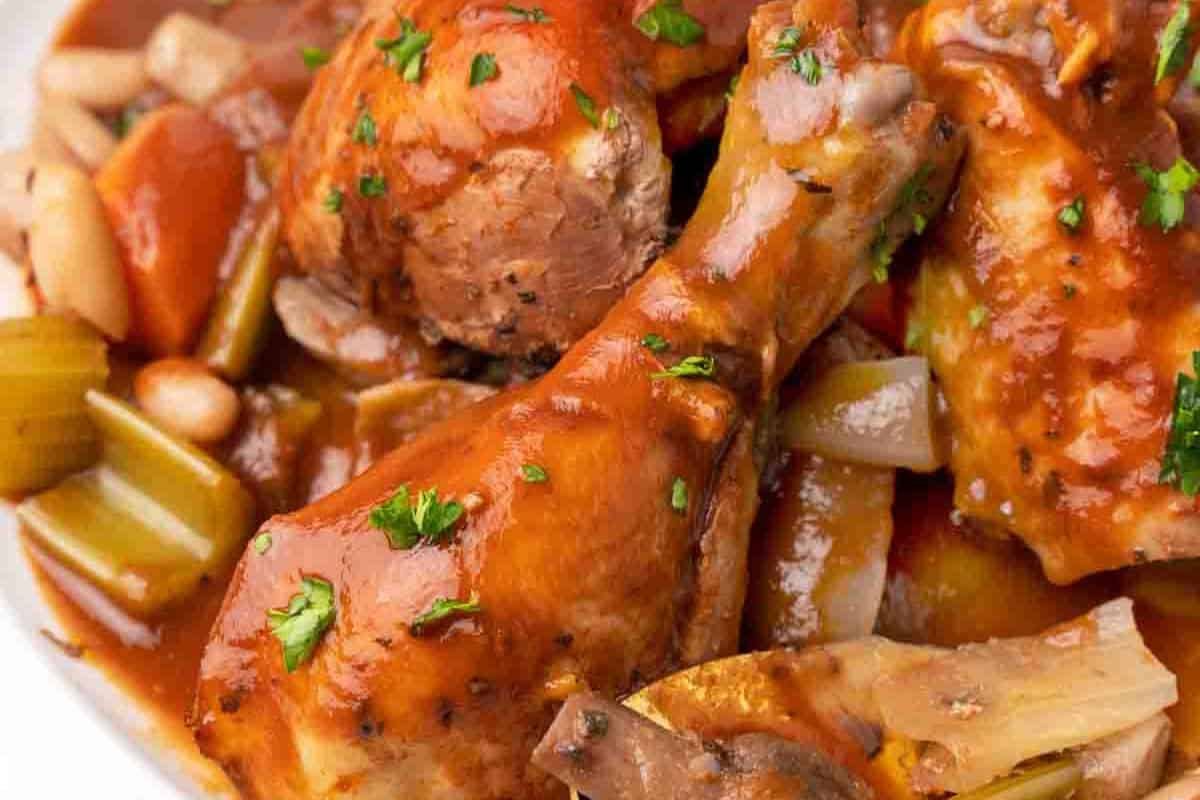 A close-up of a plate of Chicken Merlot. Chicken drumsticks and thighs are braised in a rich, reddish-brown sauce with visible carrots, celery, onions, and herbs, likely parsley.