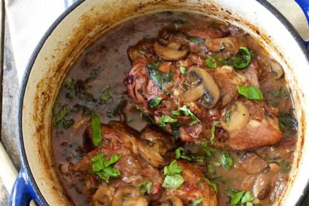 An overhead shot of a Dutch oven filled with Chicken Pinot Noir. Chicken pieces and sliced mushrooms are simmered in a dark red wine sauce and garnished with fresh basil leaves.
