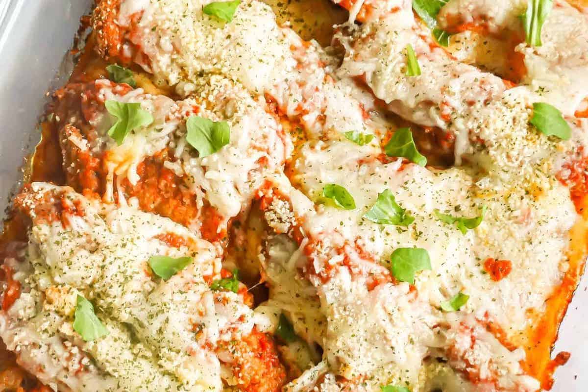 An overhead shot of a baking dish filled with Chicken Tenders Parmesan. Breaded chicken tenders are covered in red tomato sauce, melted white mozzarella cheese, and sprinkled with grated Parmesan cheese and fresh green basil leaves.