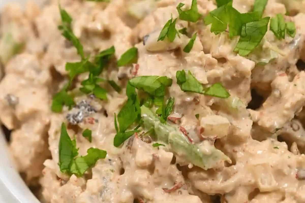 A close-up overhead shot of Chipotle Chicken Salad in a white bowl. Shredded chicken is mixed with a creamy, light brown dressing and visible pieces of red peppers and celery.