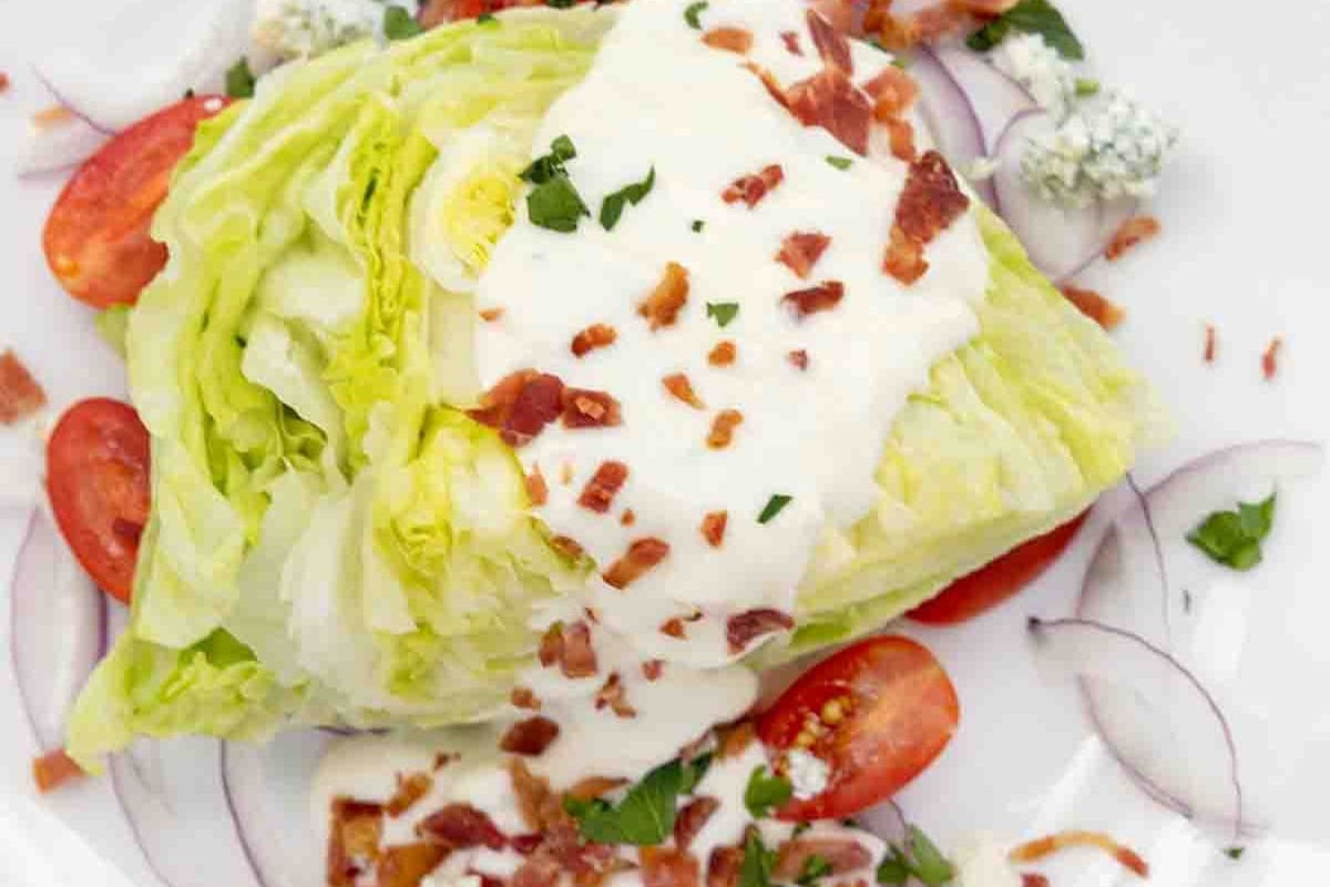 An overhead shot of a Classic Wedge Salad on a white plate. A wedge of crisp iceberg lettuce is topped with creamy white dressing, crumbled bacon, halved red tomatoes, and blue cheese crumbles.