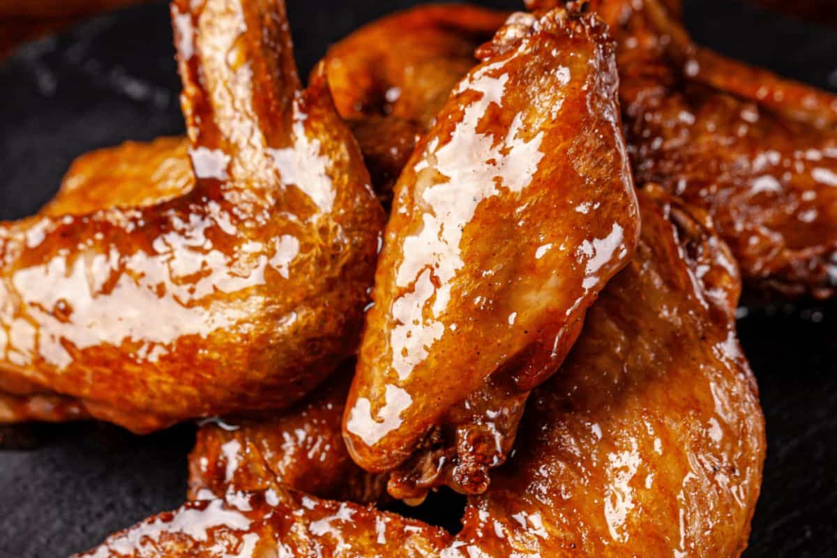 A close-up of several glistening, dark brown Coca-Cola Chicken wings on a dark surface. The wings have a sticky, glazed appearance.