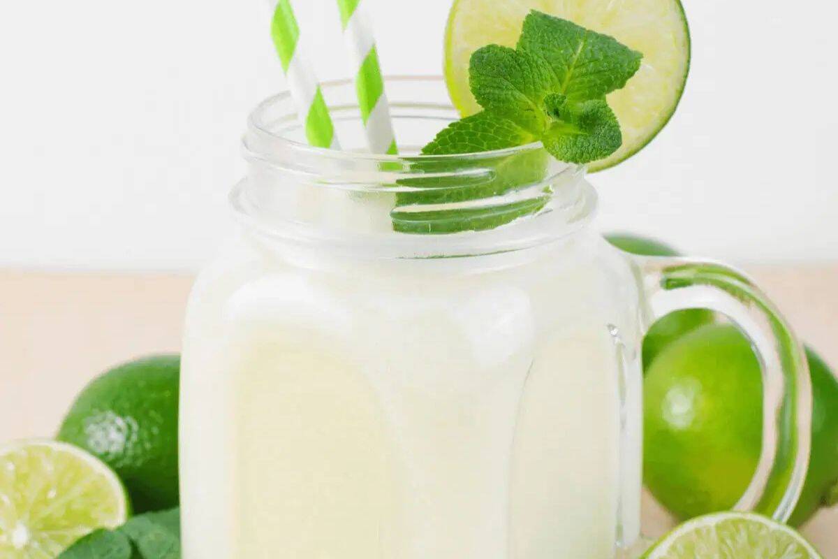  A close-up shot of a creamy white Coconut Mojito in a clear glass jar with a handle. The drink is garnished with a lime slice and a sprig of fresh mint. A striped straw is visible.