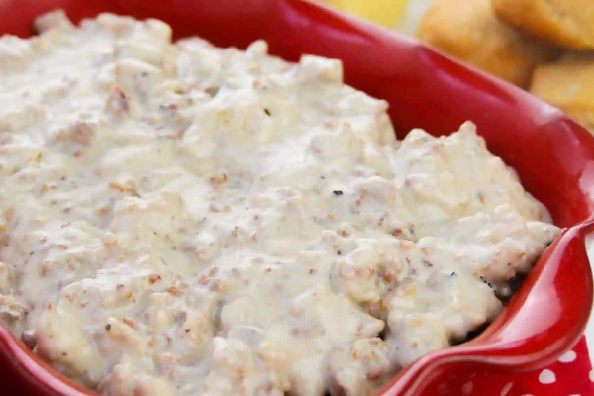 A red dish filled with creamy sausage gravy, with pieces of sausage visible; biscuits are partially seen in the background—a nod to classic recipes and timeless meals.