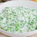 A white bowl filled with creamed peas in a creamy white sauce, placed on a light-colored tablecloth.