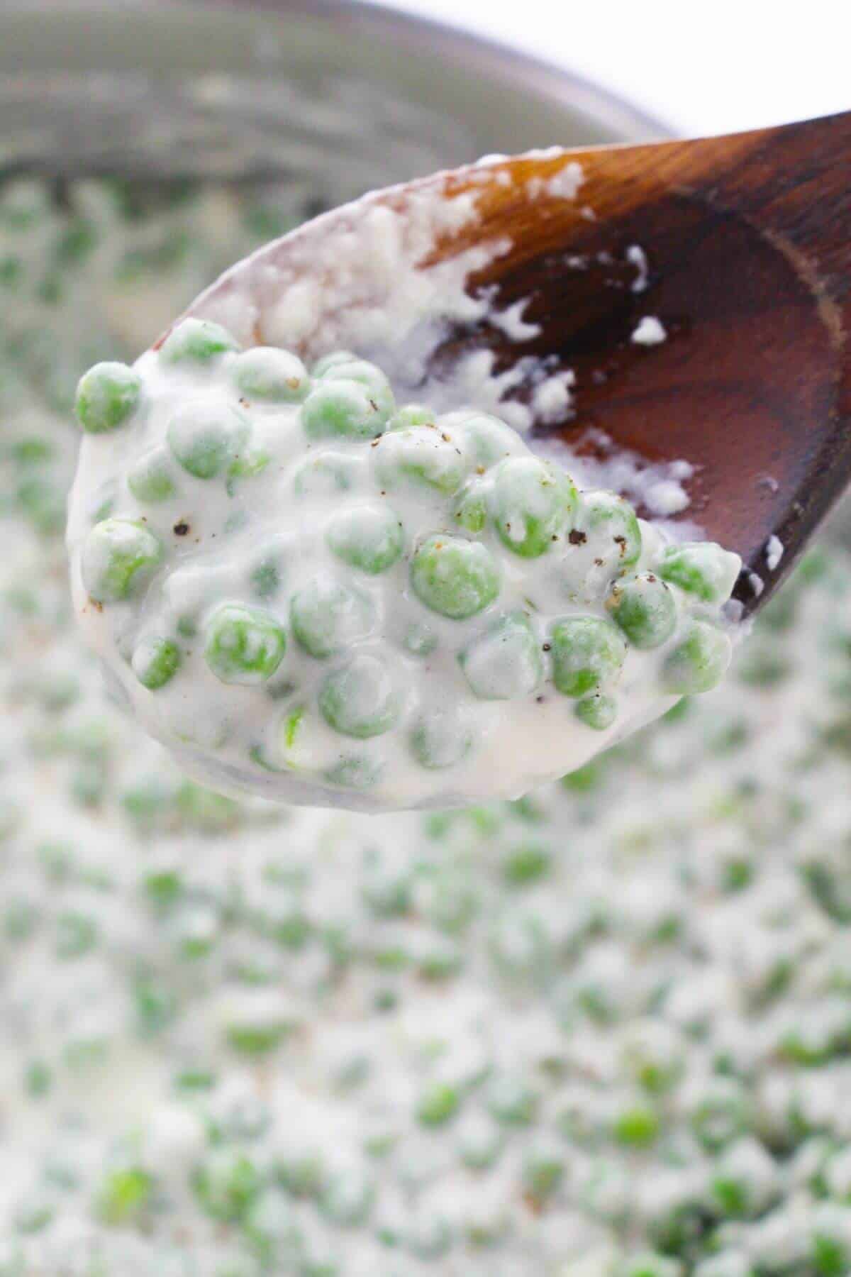 A wooden spoon holding the side above a pot filled with more creamy green peas.