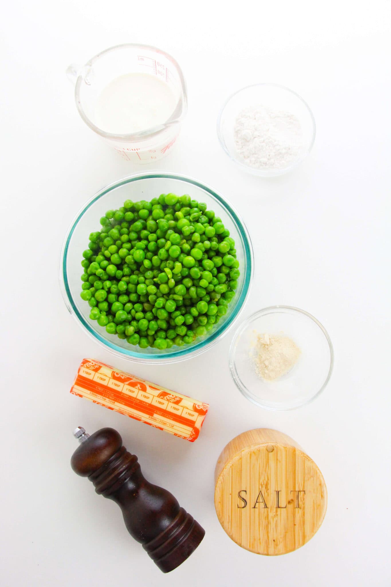 A glass bowl of green peas, butter, a pepper grinder, a salt container, flour, milk, and granulated seasoning sit on a white surface—everything you need to make classic creamed peas.