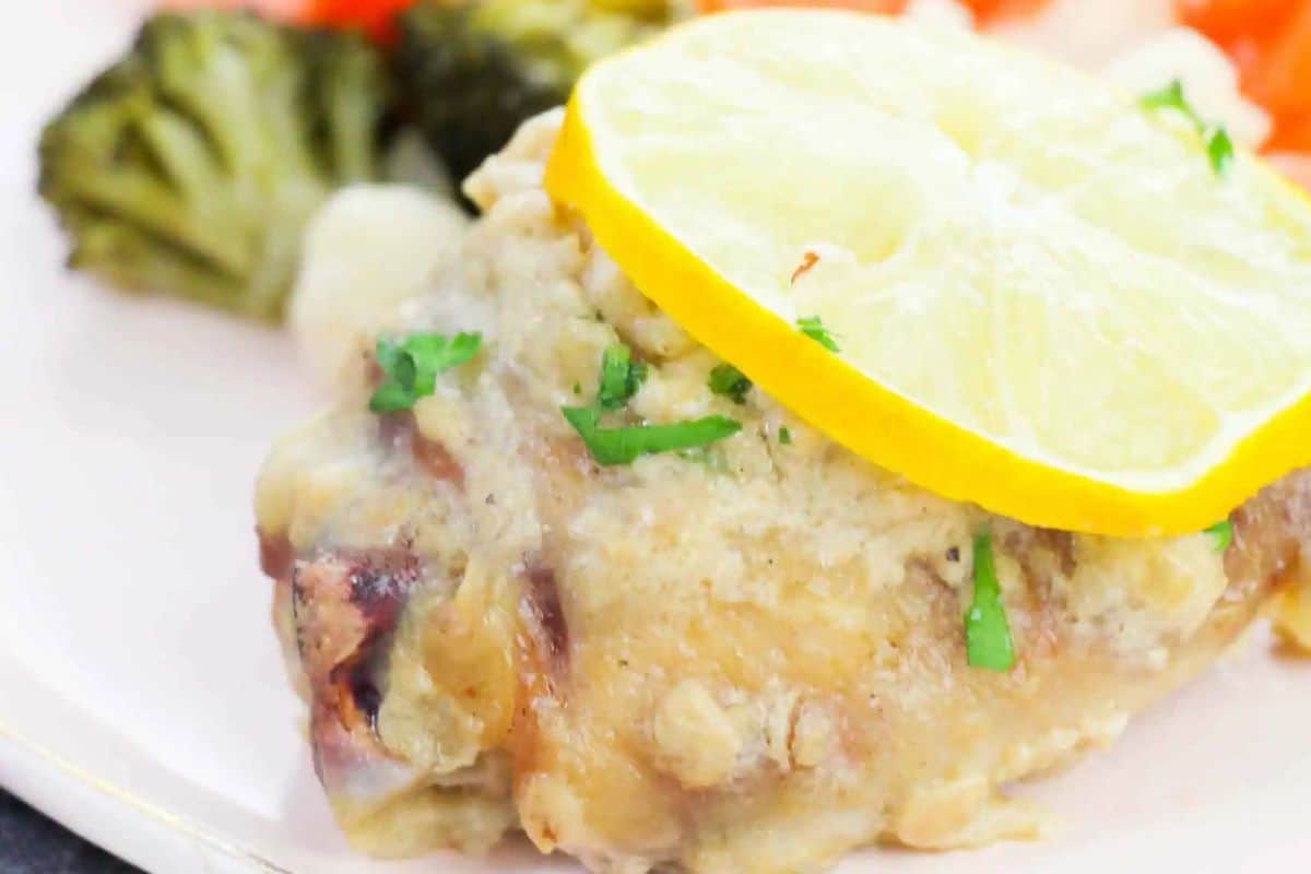 A cooked chicken thigh topped with a lemon slice and herbs, served with broccoli and carrots on a white plate—perfect for fans of juicy chicken thighs or those seeking new chicken thigh recipes.