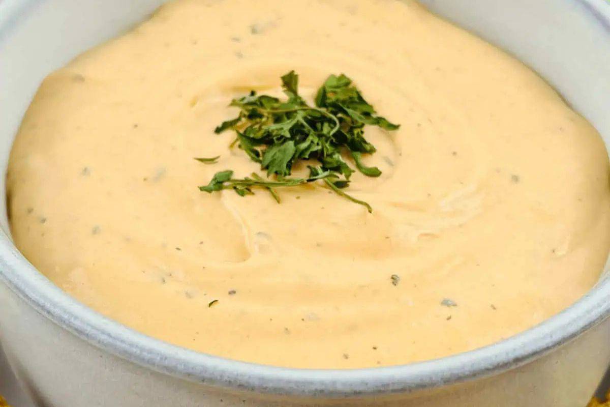 A close-up overhead shot of a white bowl filled with creamy, pale orange Buffalo Ranch Dip, garnished with fresh green parsley. Small flecks of spices are visible throughout the dip.