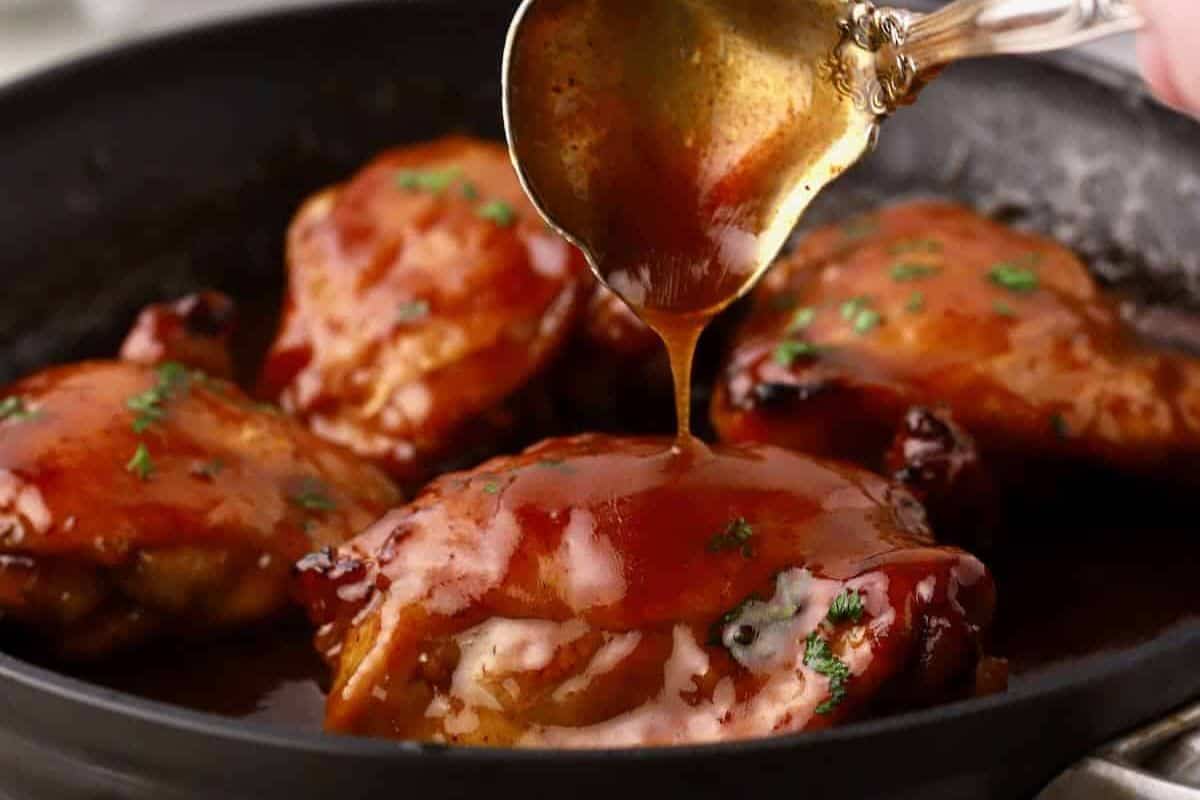 A spoon drizzles sauce over juicy chicken thighs in a skillet, with parsley sprinkled on top—a delicious addition to your favorite chicken thigh recipes.