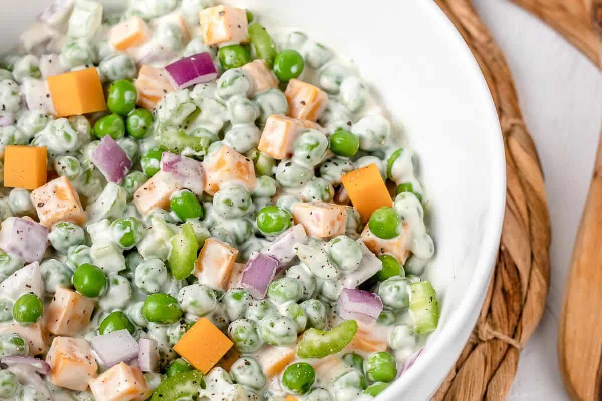 A close-up overhead shot of English Pea Salad in a white bowl. Bright green English peas are mixed with orange and white cheese cubes, diced purple red onion, and chopped light green celery, all coated in a creamy white dressing.