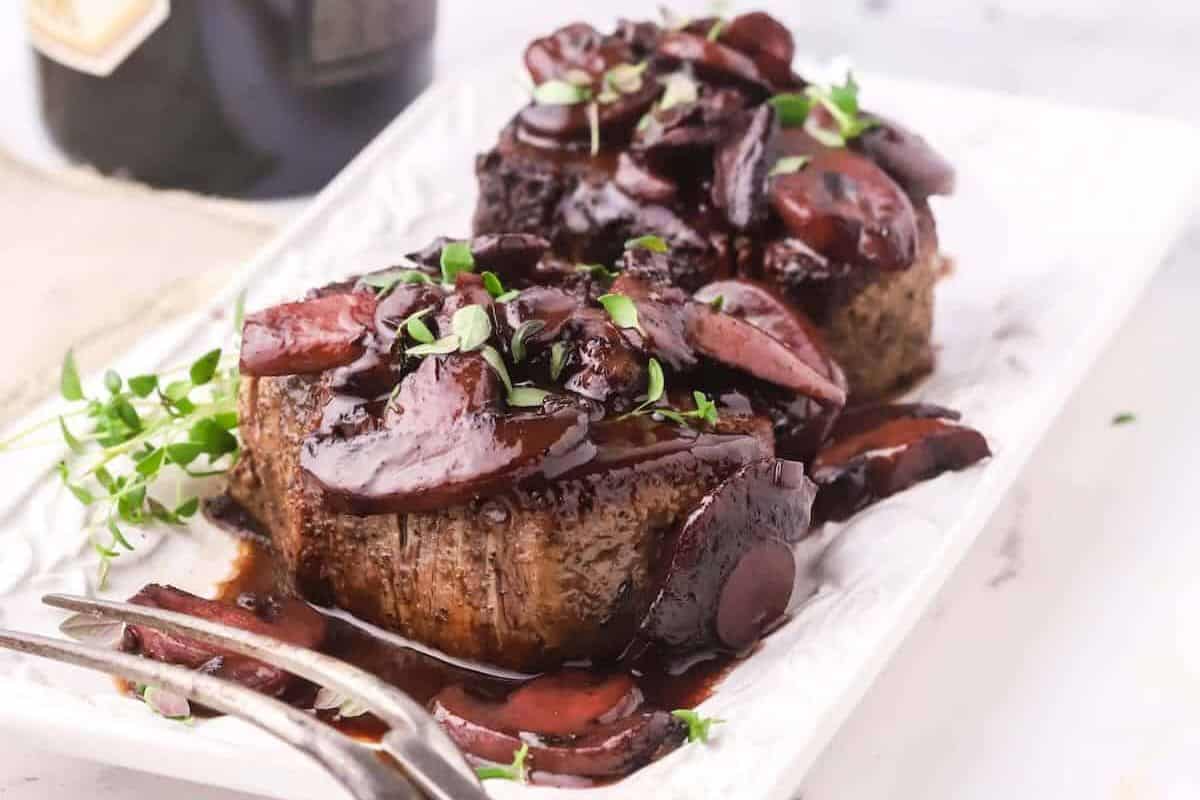 A close-up of two Filet Mignon steaks, each topped with a rich, dark red wine sauce featuring sliced mushrooms. Fresh thyme sprigs are used as a garnish.
