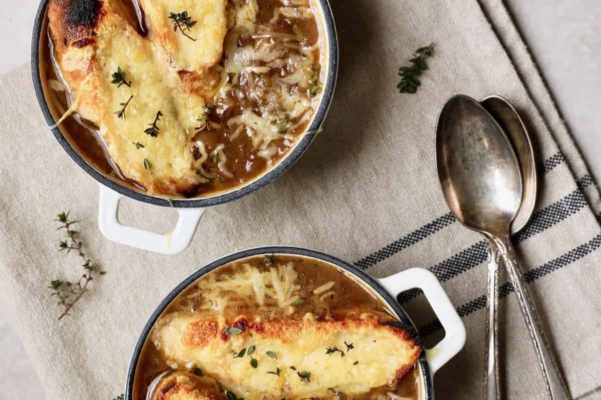Two bowls of delicious French onion soup, perfect for two, are topped with toasted bread and melted cheese, garnished with thyme. Two spoons rest on the napkin beside the bowls.