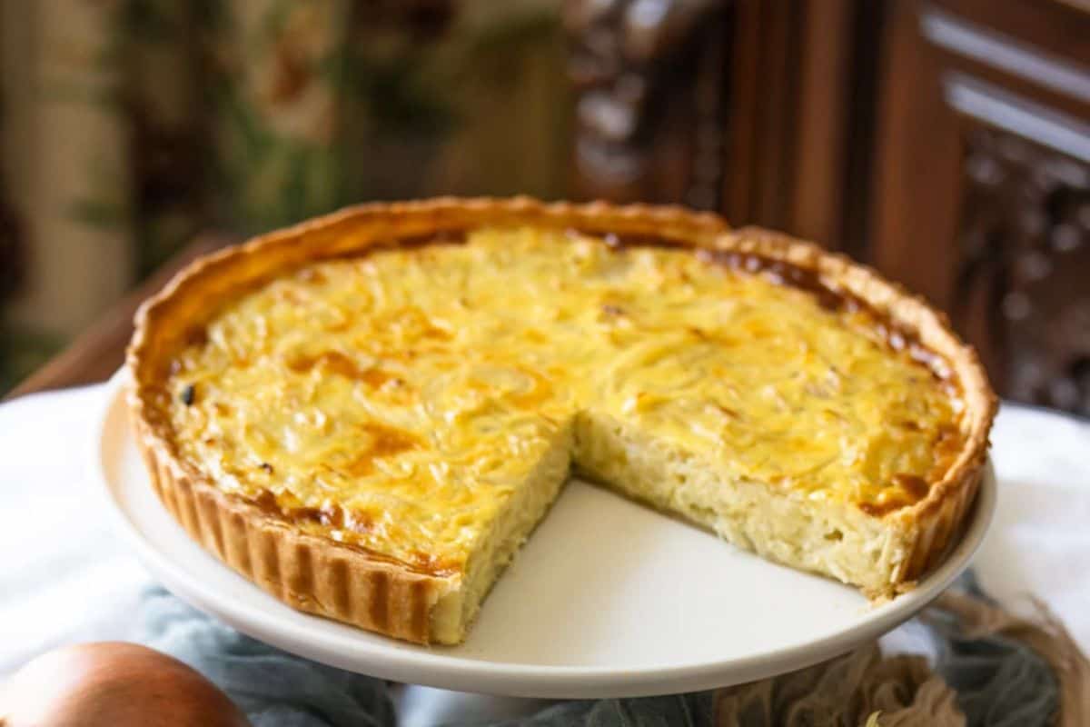 A round onion tart with a slice missing sits on a white plate, showing its golden-brown crust and creamy filling—a nod to vintage dishes and timeless meals.