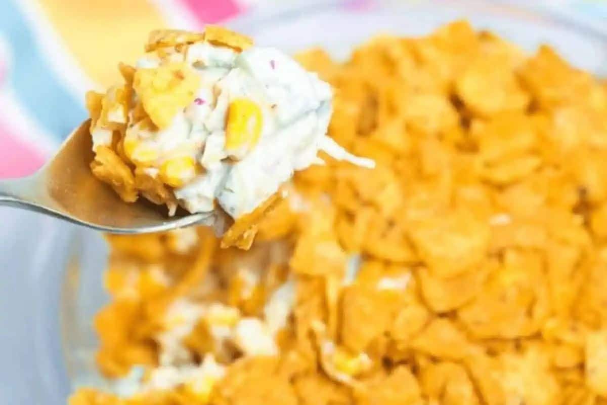 A close-up of a spoonful of Frito Corn Salad being lifted from a glass dish. The salad features crushed golden Frito corn chips, white creamy dressing, and yellow corn kernels.