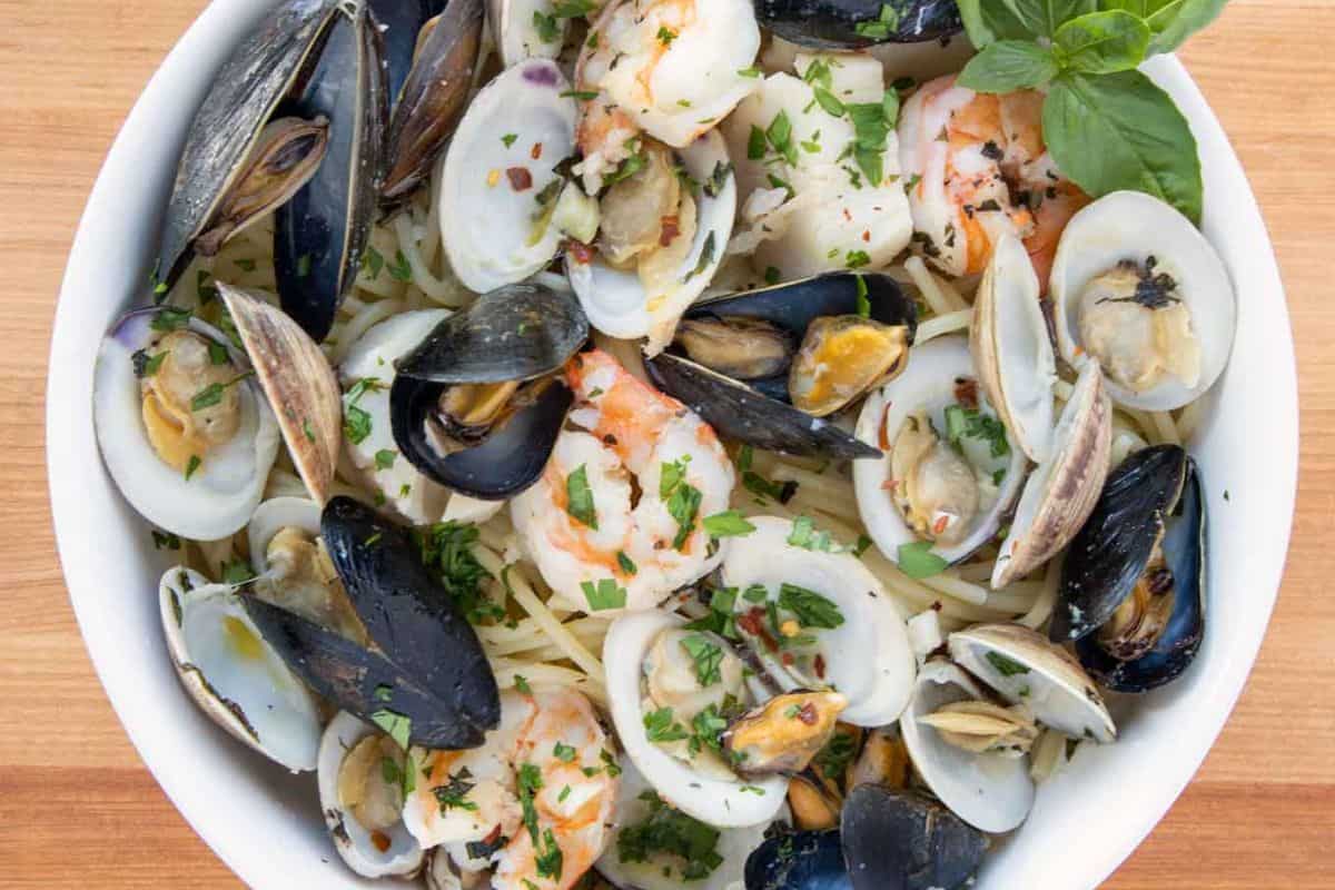 An overhead shot of a white bowl filled with Frutti di Mare. Spaghetti is tossed with a generous mix of mussels, clams, and shrimp, seasoned with herbs and spices. A sprig of fresh basil garnishes the dish.