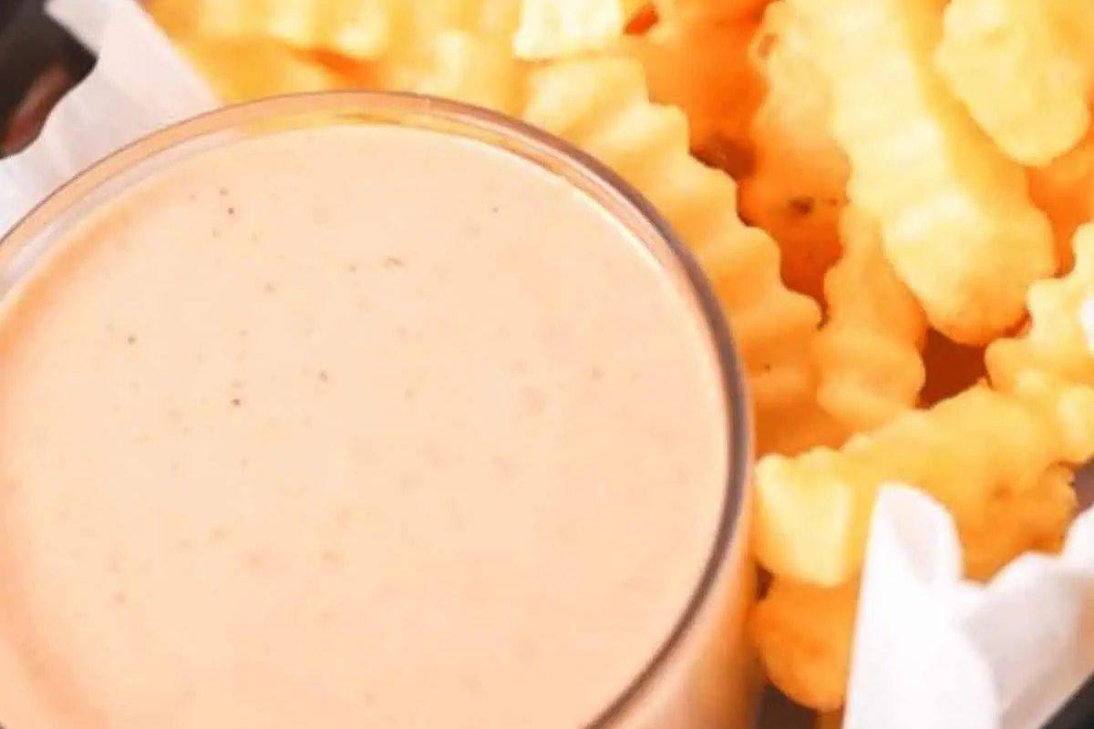 A close-up overhead shot of a clear glass filled with creamy, pale orange-pink Fry Sauce, next to a serving of golden-brown crinkle-cut french fries.