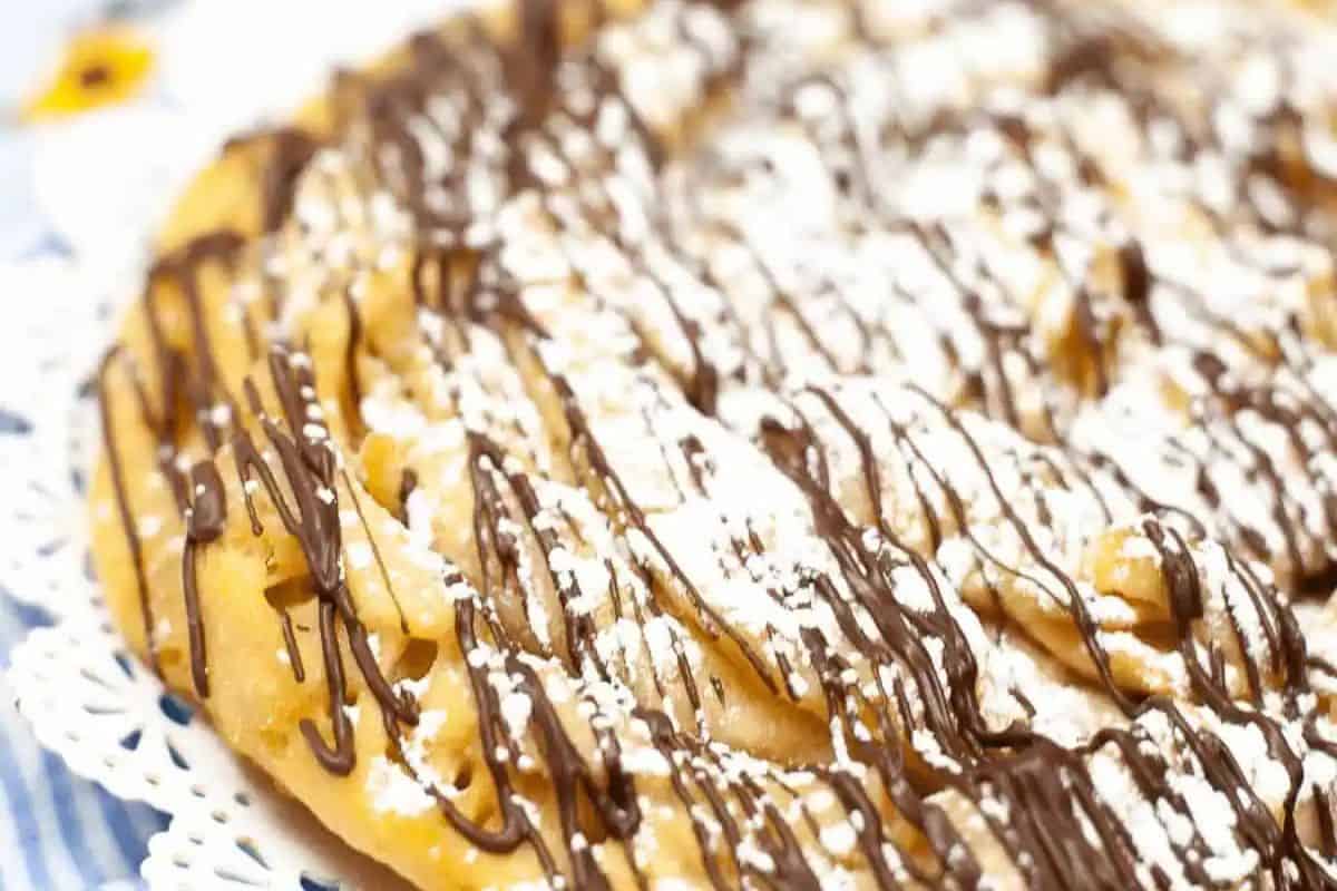 A close-up of a large funnel cake topped with powdered sugar and drizzled with chocolate on a doily—a game changing treat that elevates any dessert table.