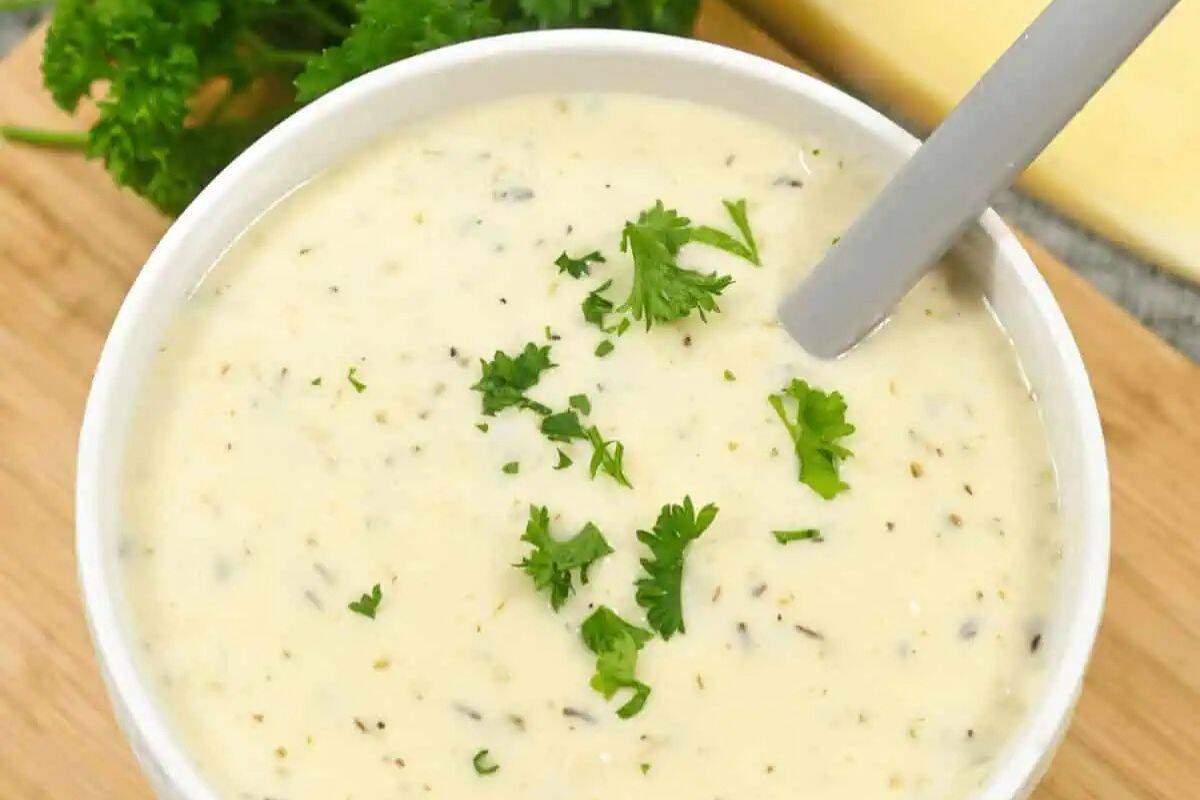 A close-up overhead shot of a white bowl filled with creamy, pale white Garlic Parmesan Sauce. Small flecks of herbs and possibly grated Parmesan are visible throughout the sauce, which is garnished with fresh green parsley leaves.