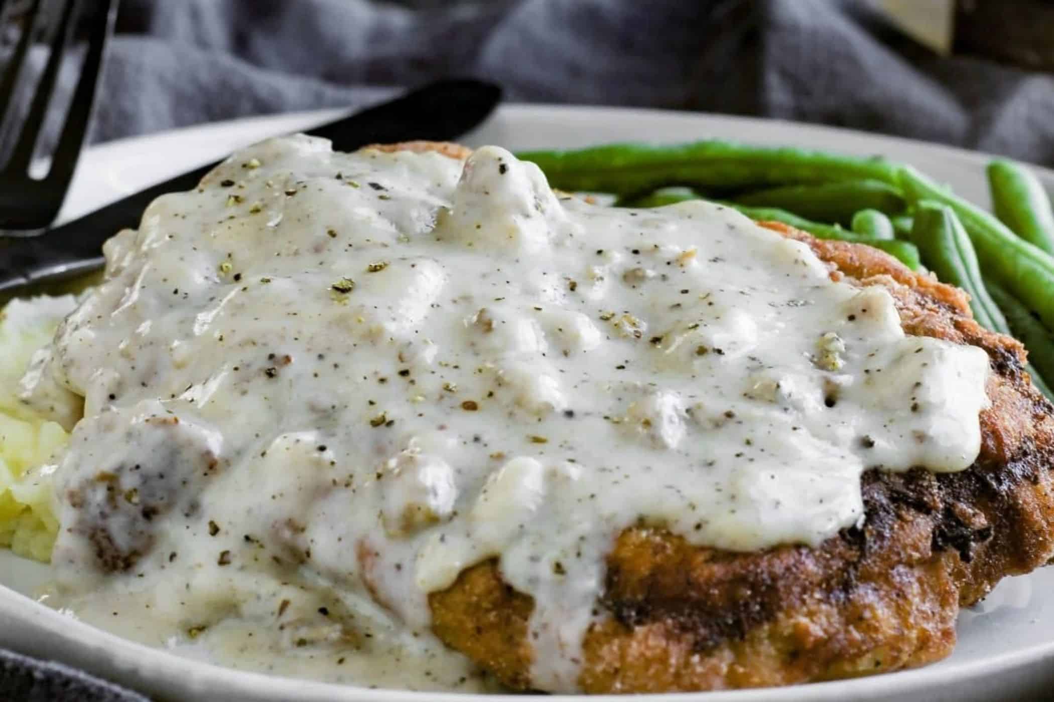 A gluten free breaded cube steak covered in white pepper gravy on a plate with mashed potatoes and green beans—perfect for busy weeknights and anyone seeking easy cube steak recipes.