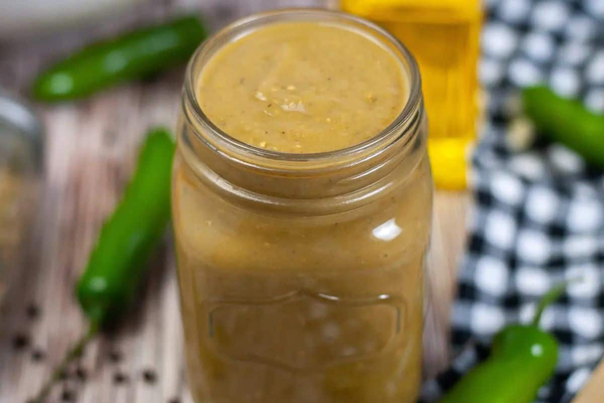  A clear glass jar filled with a smooth, pale green Green Chili Enchilada Sauce. Several fresh green chili peppers are scattered around the jar on a wooden surface.
