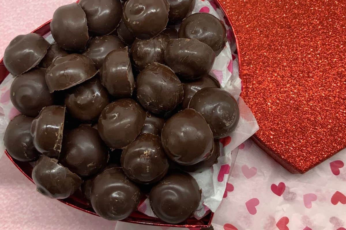 A heart-shaped box filled with round chocolate caramel candies, perfect for indulgent desserts, placed on heart-patterned tissue paper next to a red glittery heart-shaped lid.