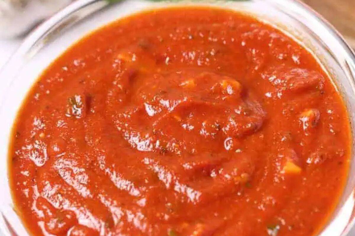 A close-up overhead shot of a clear glass bowl filled with rich, vibrant red Homemade Pizza Sauce. Visible chunks of tomatoes and herbs are present throughout the sauce.