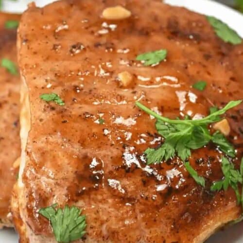 Close-up of a delicious, glazed grilled pork chop garnished with cilantro and small garlic pieces on a white plate, perfect for two.