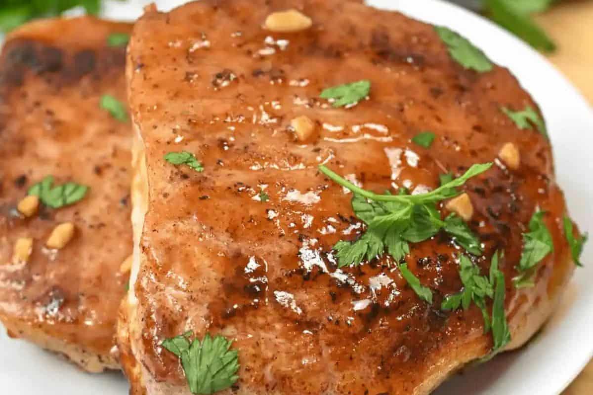 Close-up of a delicious, glazed grilled pork chop garnished with cilantro and small garlic pieces on a white plate, perfect for two.