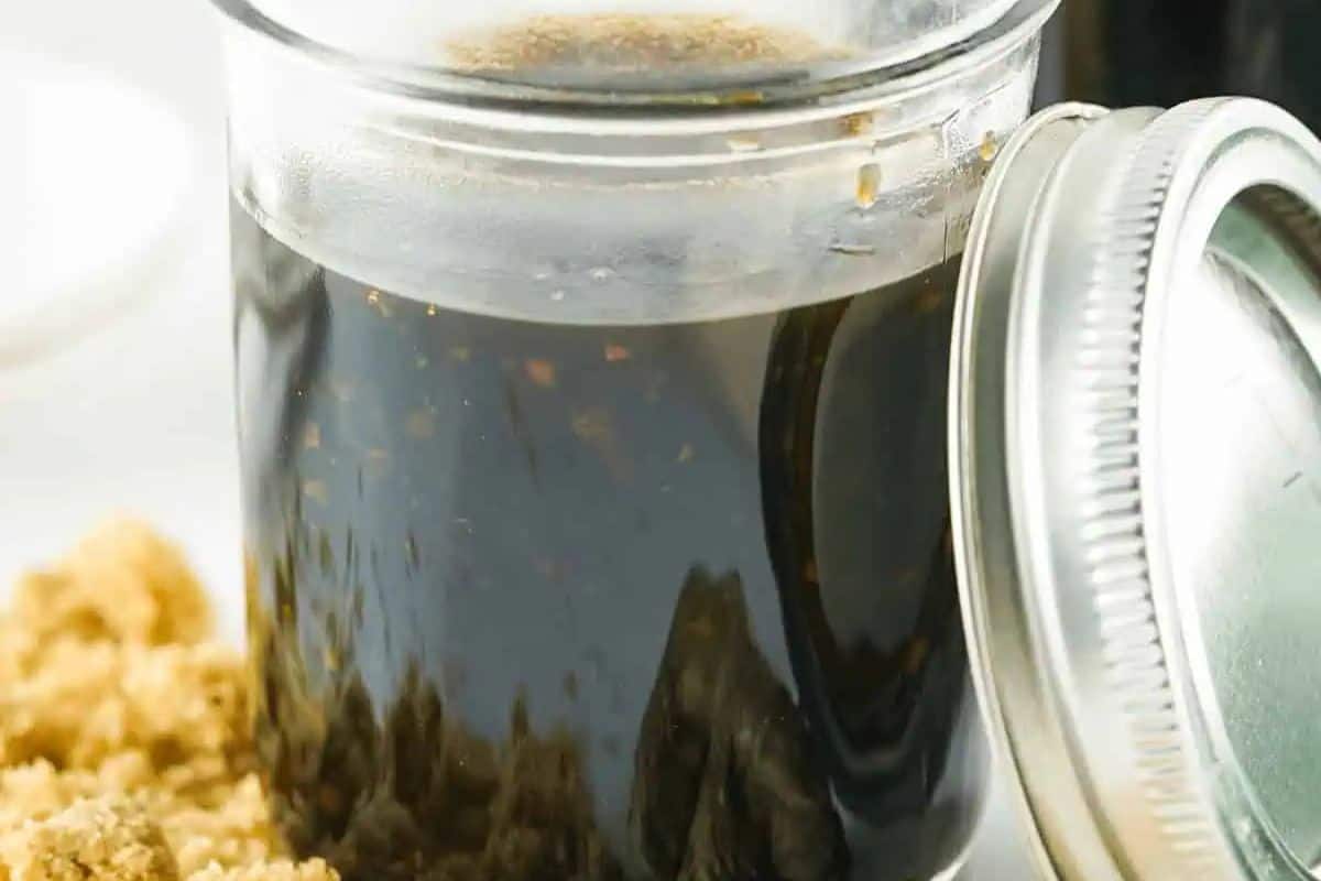 A close-up of a clear glass jar filled with dark brown Honey Teriyaki Sauce. Some brown sugar is visible near the jar.