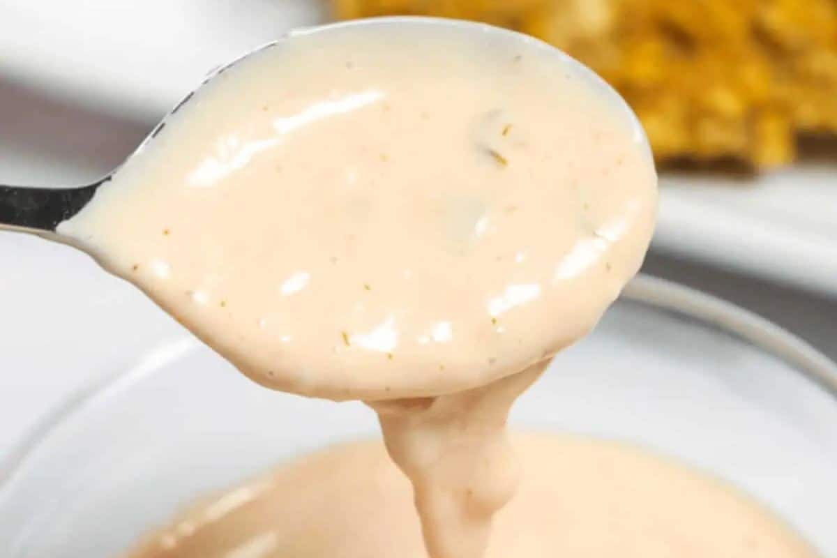 A close-up shot of a spoonful of creamy, pale orange-pink In-N-Out Sauce dripping back into a bowl filled with more of the sauce. Small relish-like chunks are visible throughout.