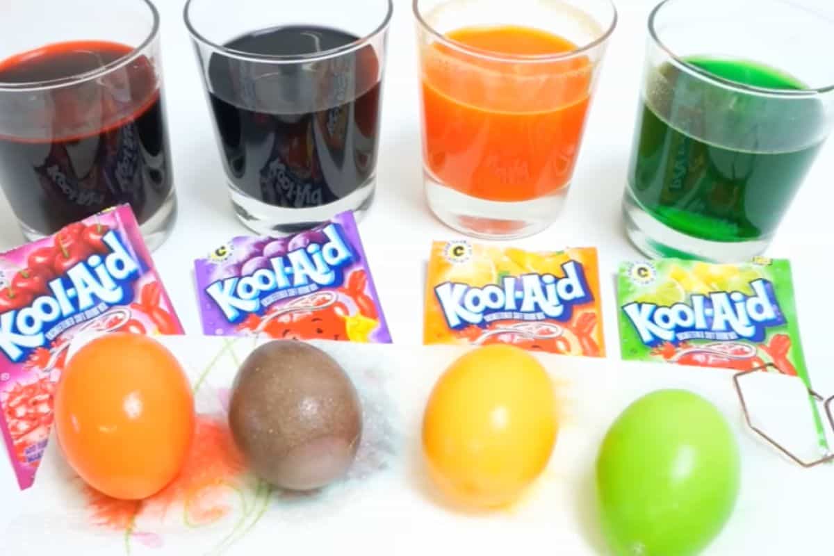 Four dyed eggs in orange, purple, yellow, and green sit in front of four glasses of colored liquid and Kool-Aid packets matching each color—perfect for easy Easter egg decorating or creative Easter egg ideas.