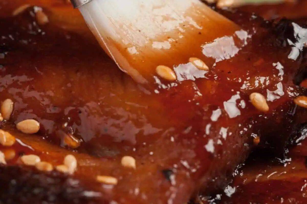 A close-up shot of a brush applying thick, glossy, dark reddish-brown Korean BBQ Sauce onto ribs or meat, with visible sesame seeds.
