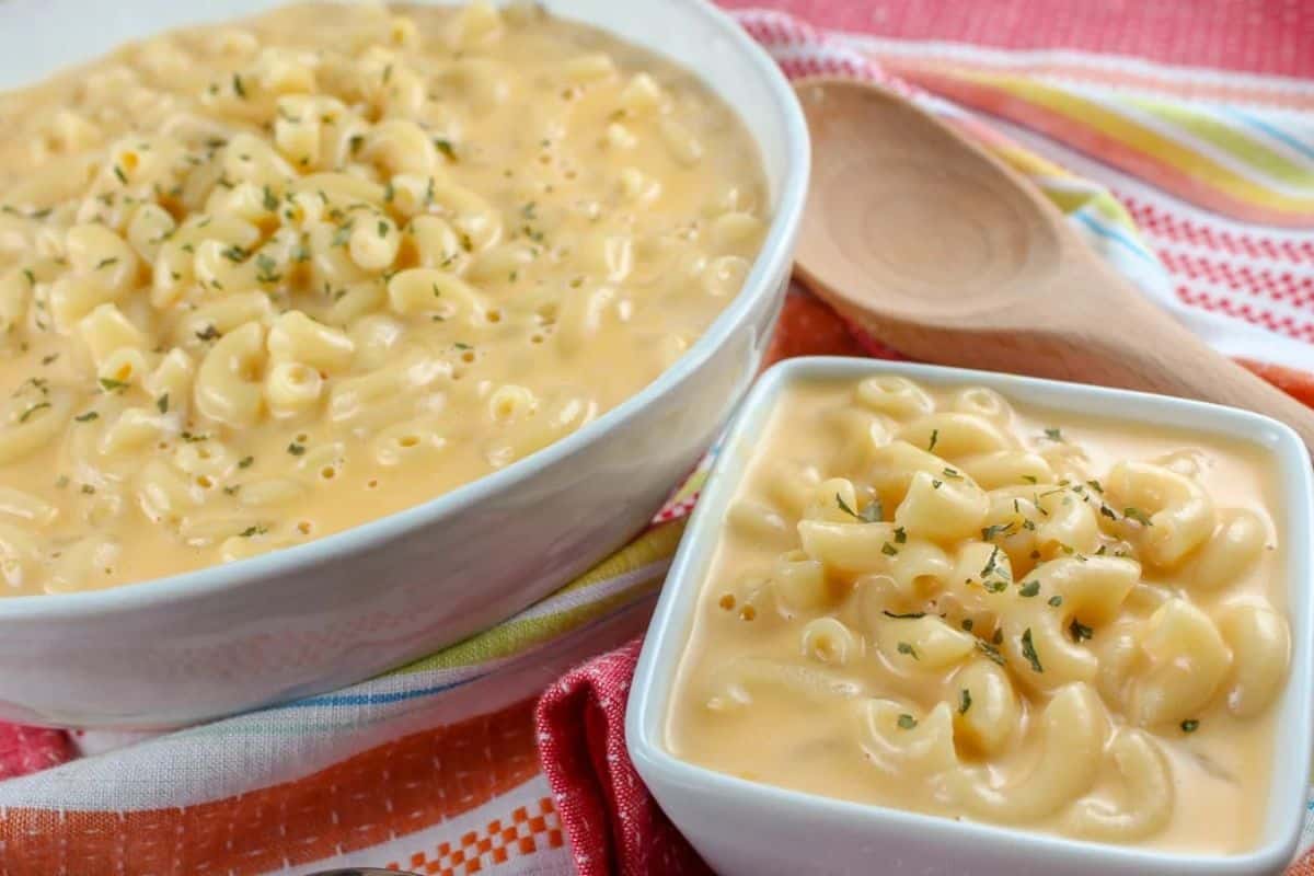 Elbow macaroni pasta is coated in a smooth, creamy, bright yellow cheese sauce and sprinkled with chopped green herbs. A wooden spoon rests near the larger bowl.