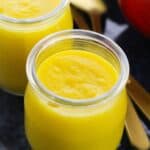 Two small glass jars filled with smooth Mango Pudding sit beside gold-colored spoons on a dark surface.