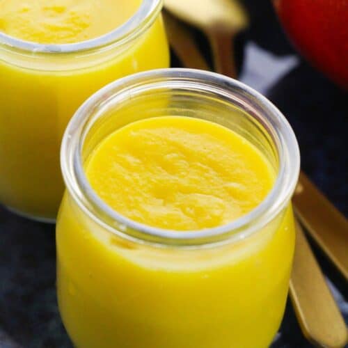Two small glass jars filled with smooth Mango Pudding sit beside gold-colored spoons on a dark surface.