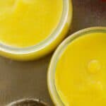 Two small glass jars filled with smooth, bright yellow mango pudding are placed on a metallic surface next to a decorative object.