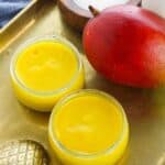 Two small glass jars filled with bright yellow mango pudding sit on a gold tray next to a whole mango, a wooden bowl of sugar, and a jar.