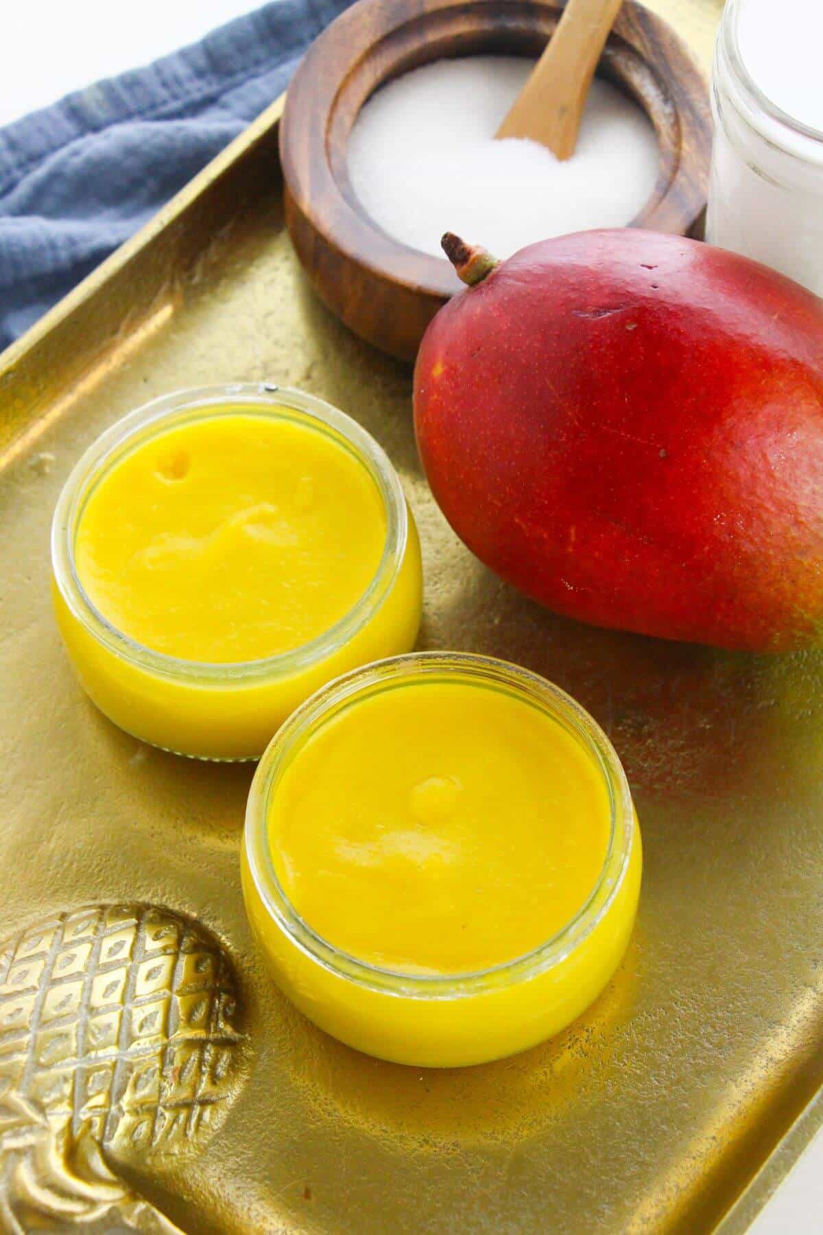 Two small glass jars filled with bright yellow mango pudding sit on a gold tray next to a whole mango, a wooden bowl of sugar, and a jar.