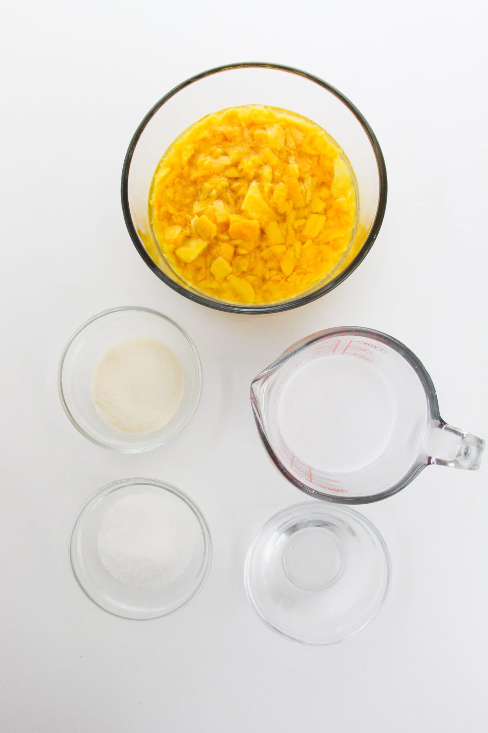 Four glass containers hold ingredients for Mango Pudding: chopped mango, sugar, water, gelatin powder, and another clear liquid, all arranged neatly on a white surface.