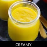 Two small glass jars filled with creamy Mango Pudding are shown on a dark surface, accompanied by two gold spoons.