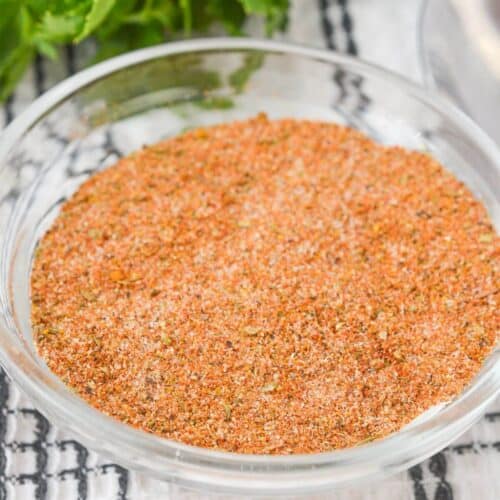 A clear glass bowl filled with meatloaf seasoning sits on a white and black checkered cloth, with fresh green herbs partially visible in the background.