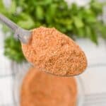 A spoonful of brownish meatloaf seasoning is held over a bowl, with a green leafy herb in the background.
