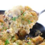 A close-up of a serving spoon lifting a portion topped with crispy potatoes garnished with chopped parsley, from a skillet.