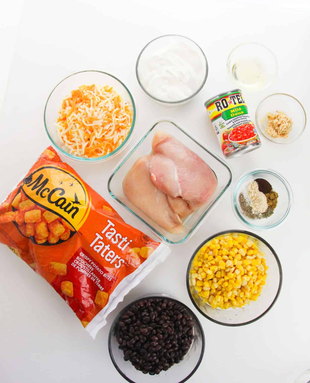 A flat lay of Chicken Tater Tot Casserole ingredients: raw chicken breasts, shredded cheese, sour cream, canned corn, black beans, Rotel tomatoes, spices, minced garlic, oil, and a bag of Tasti Taters.