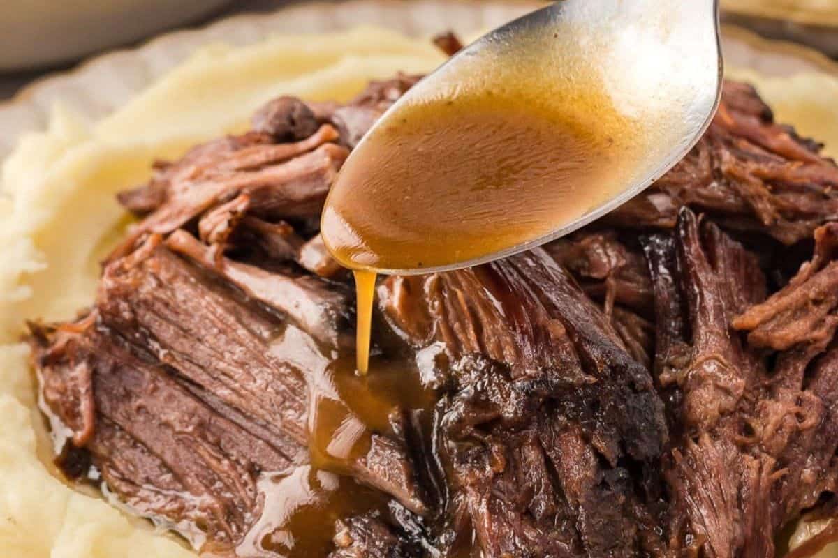 A close-up of a spoonful of rich brown gravy being poured over tender, shredded Mississippi Pot Roast, which is served on a bed of creamy mashed potatoes. 
