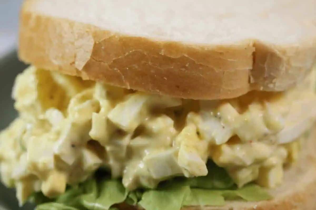 A close-up of an Egg Salad Sandwich. Creamy yellow egg salad with visible chunks of egg white and yolk is piled between two slices of white bread, resting on a bed of green lettuce.