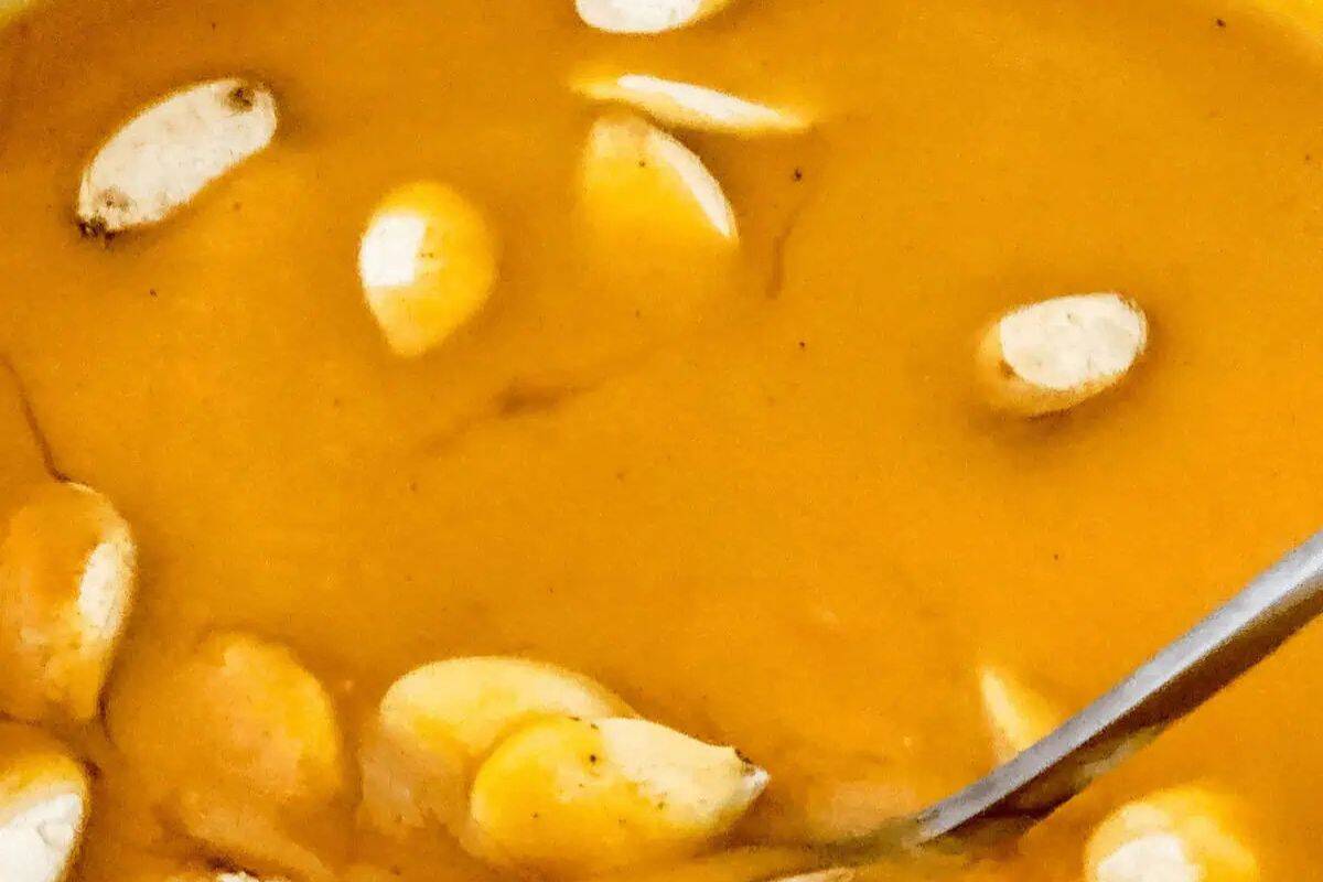Close-up of a creamy orange soup with several seeds floating on the surface, reminiscent of restaurant-inspired recipes. A metal spoon is partially visible on the right edge.