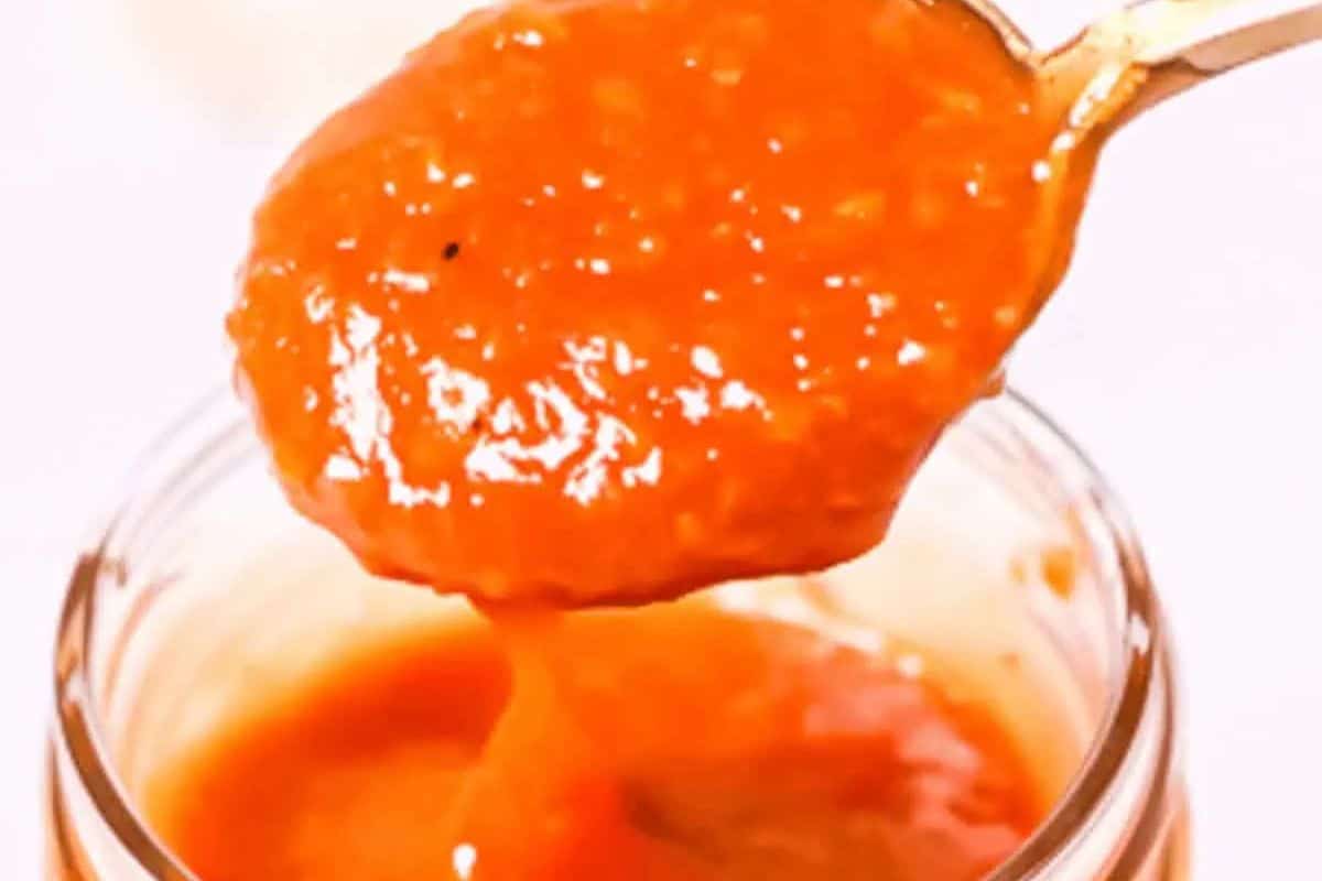  A close-up shot of a spoonful of thick, bright orange Peach BBQ Sauce being lifted from a glass jar. Visible chunks of peach are present in the sauce.